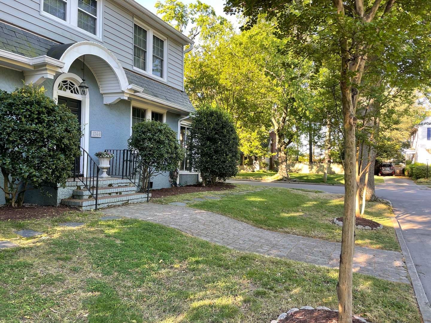 Blue house with front porch, gravel path, and green lawn. Trees line the right side near the street.
