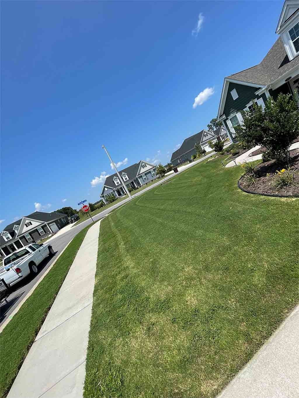 Lush green lawn with sidewalk, houses in background under bright blue sky. A white truck parked on the street.