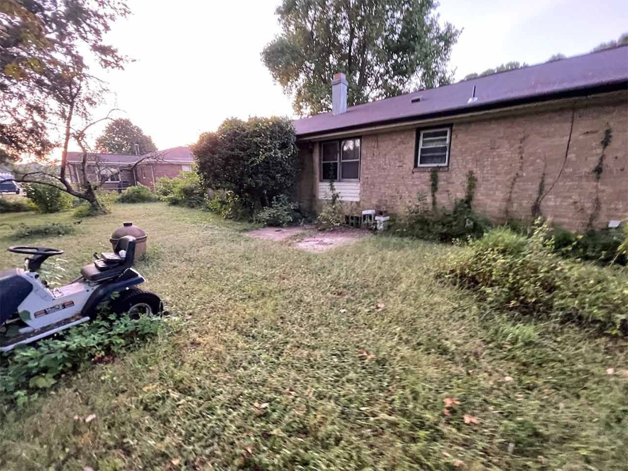 Overgrown backyard with a brick house, weeds, and a riding lawnmower.