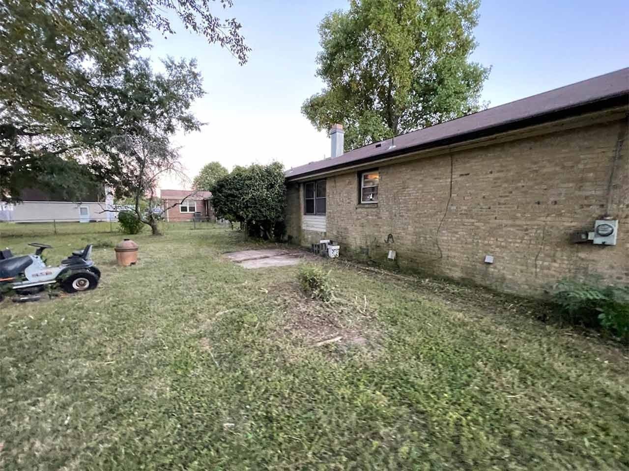 Backyard view with lawnmower, brick house, and trees under a partly cloudy sky.