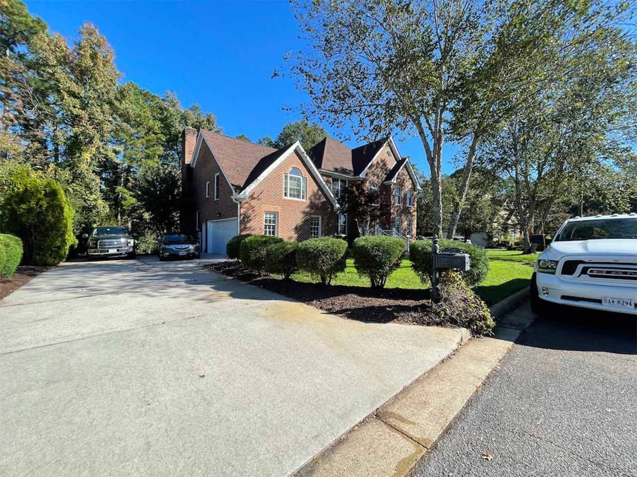 Two-story brick house with a brown roof and a concrete driveway. Trees and shrubs surround the home on a sunny day.