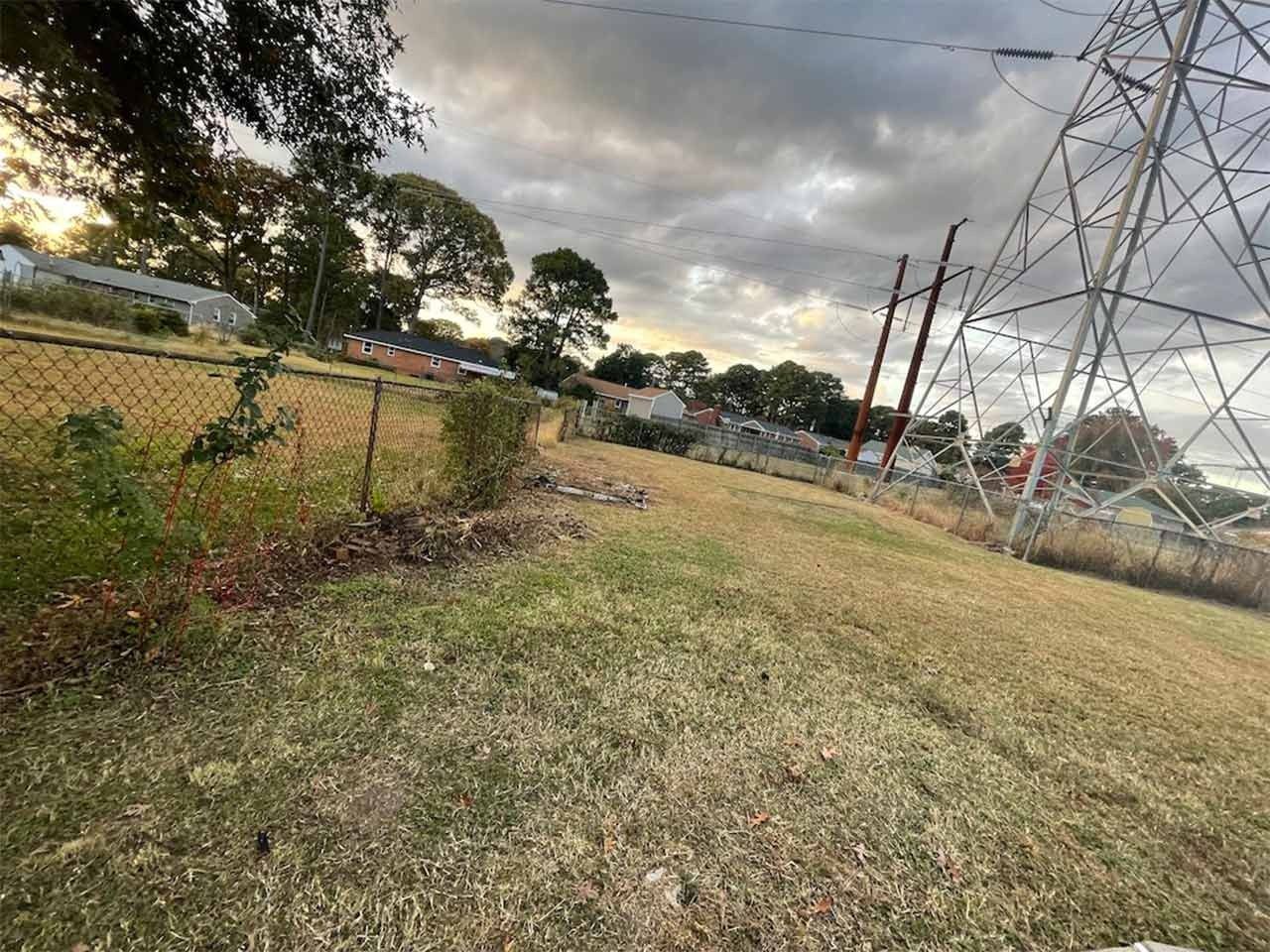 Grassy hill with chain-link fence, power lines, houses, and cloudy sky in background.