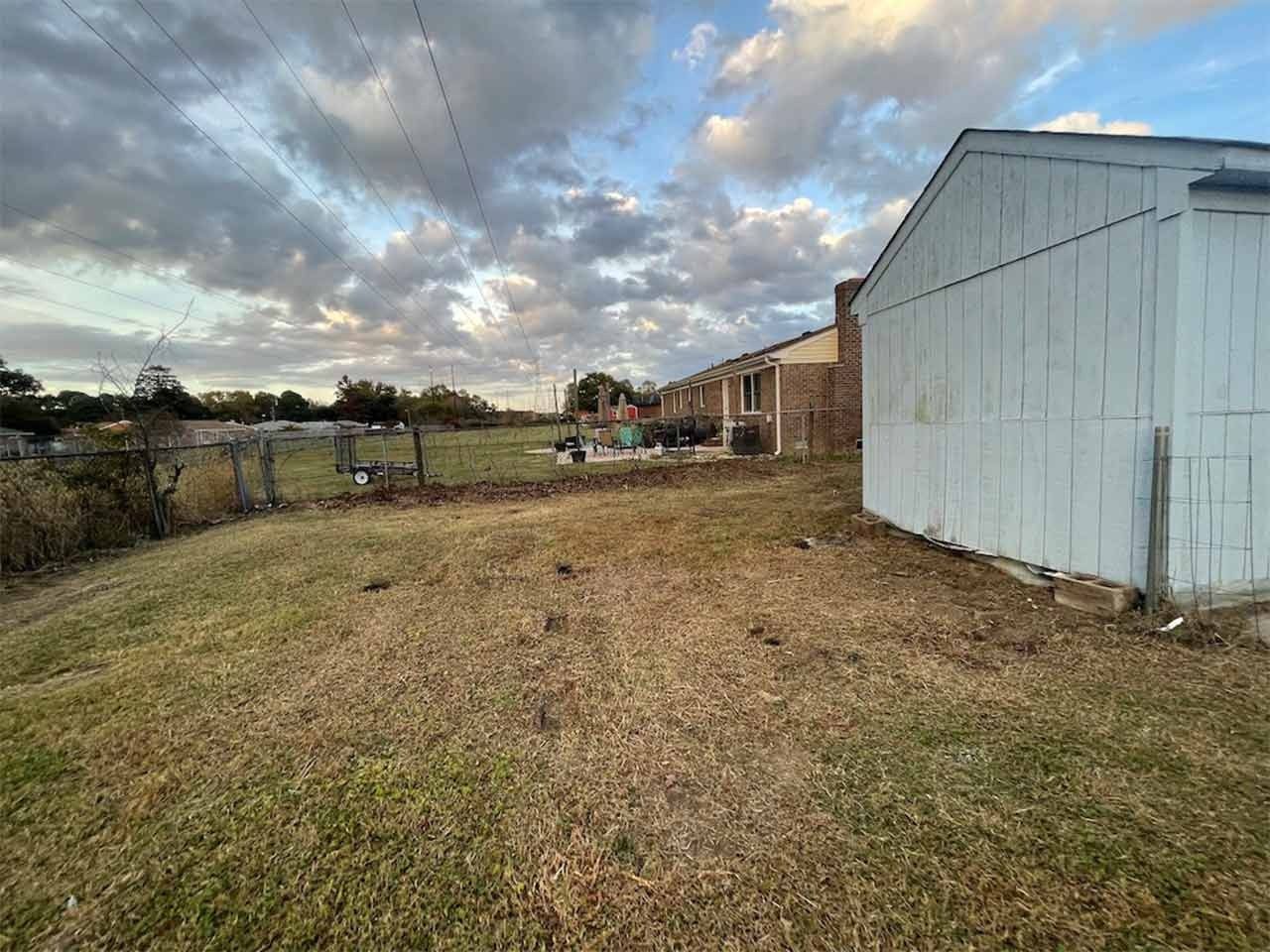 Grassy yard next to white shed. Fenced area and distant buildings under cloudy sky.