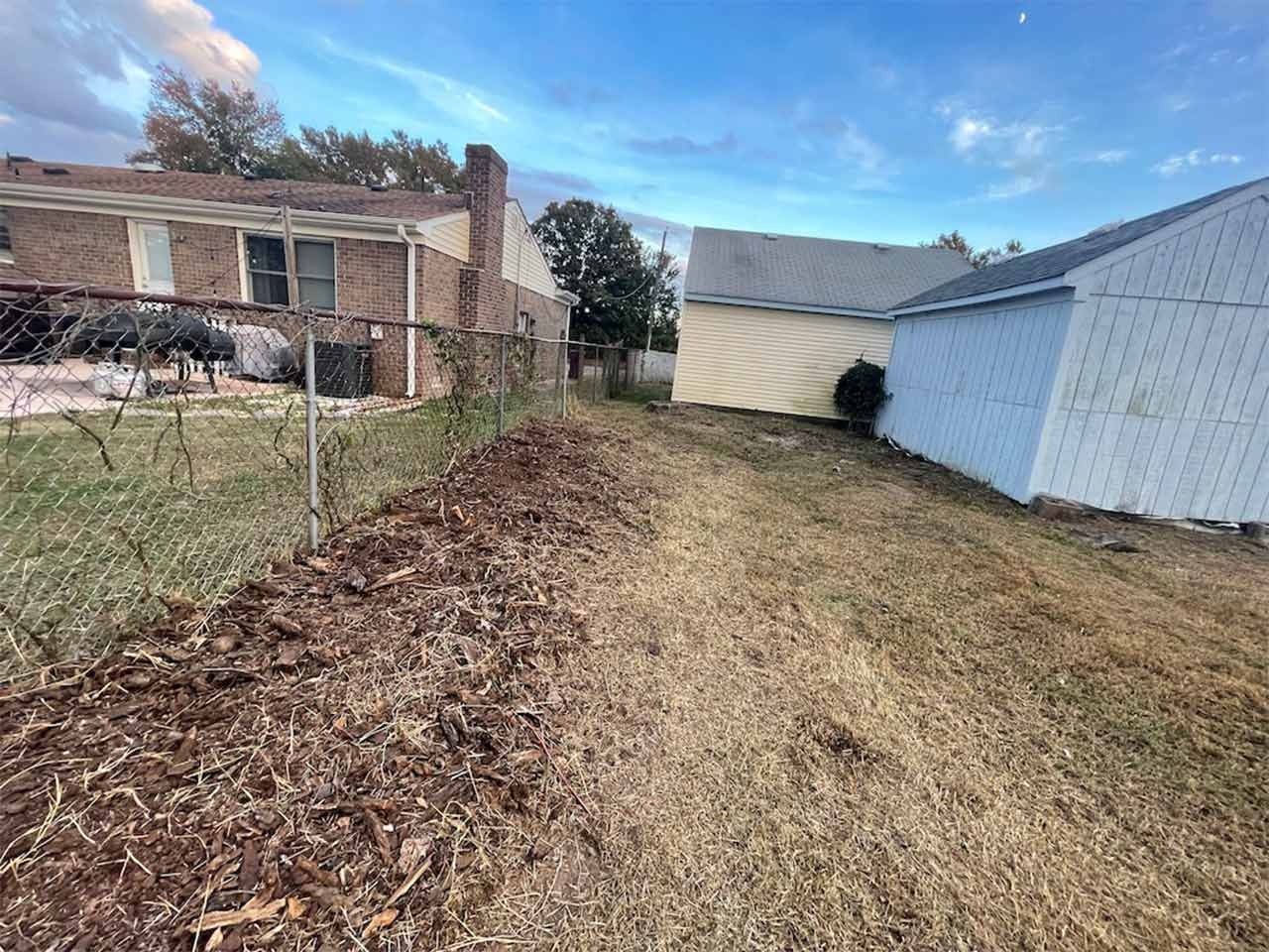 Backyard view: Chain link fence, dry grass, sheds, brick house with blue sky background.