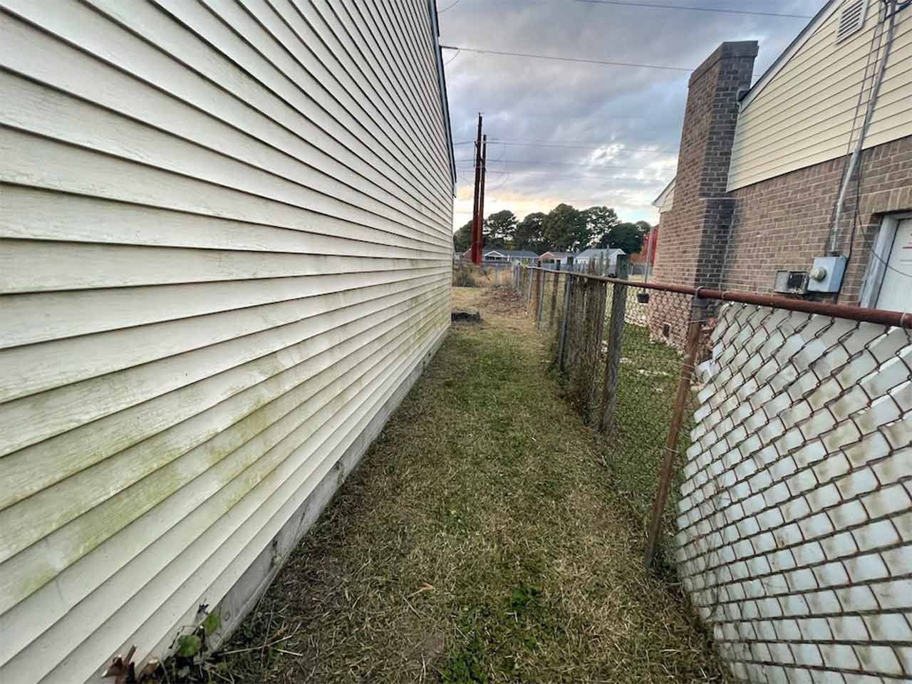 Narrow grassy passage between a white-sided building and a chain-link fence, overcast sky in background.