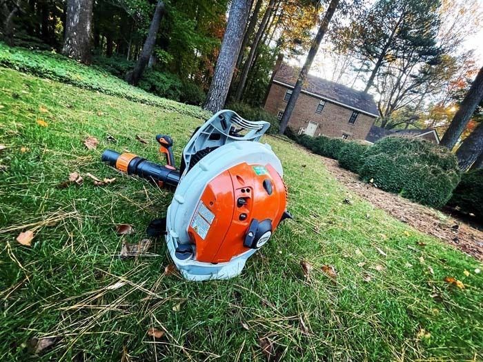 Orange and white backpack leaf blower on green grass with a house in the background.