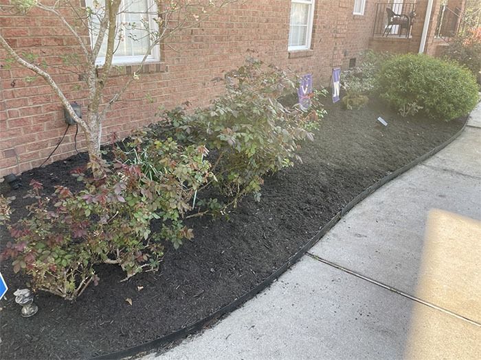 Mulched flower bed along a brick building and sidewalk; shrubs and a small tree are present.