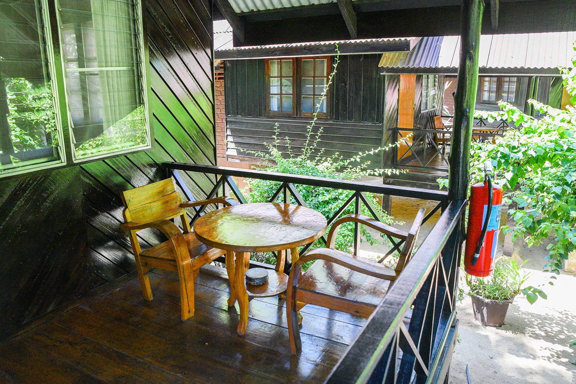 Covered porch with table and chairs overlooking a courtyard with other buildings, greenery, and a fire extinguisher.