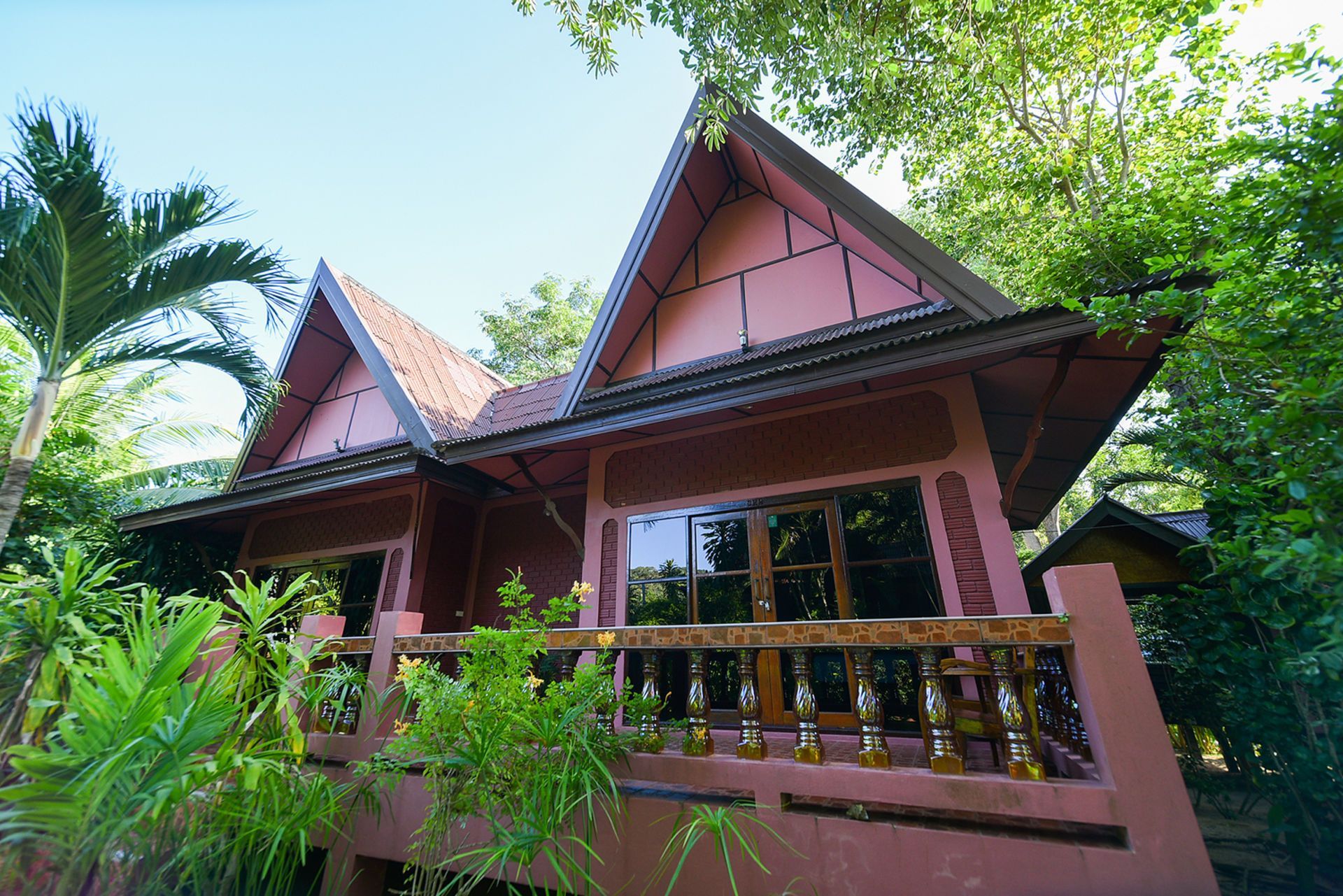 Red Thai-style bungalows with a wooden balcony surrounded by lush greenery under a sunny sky.