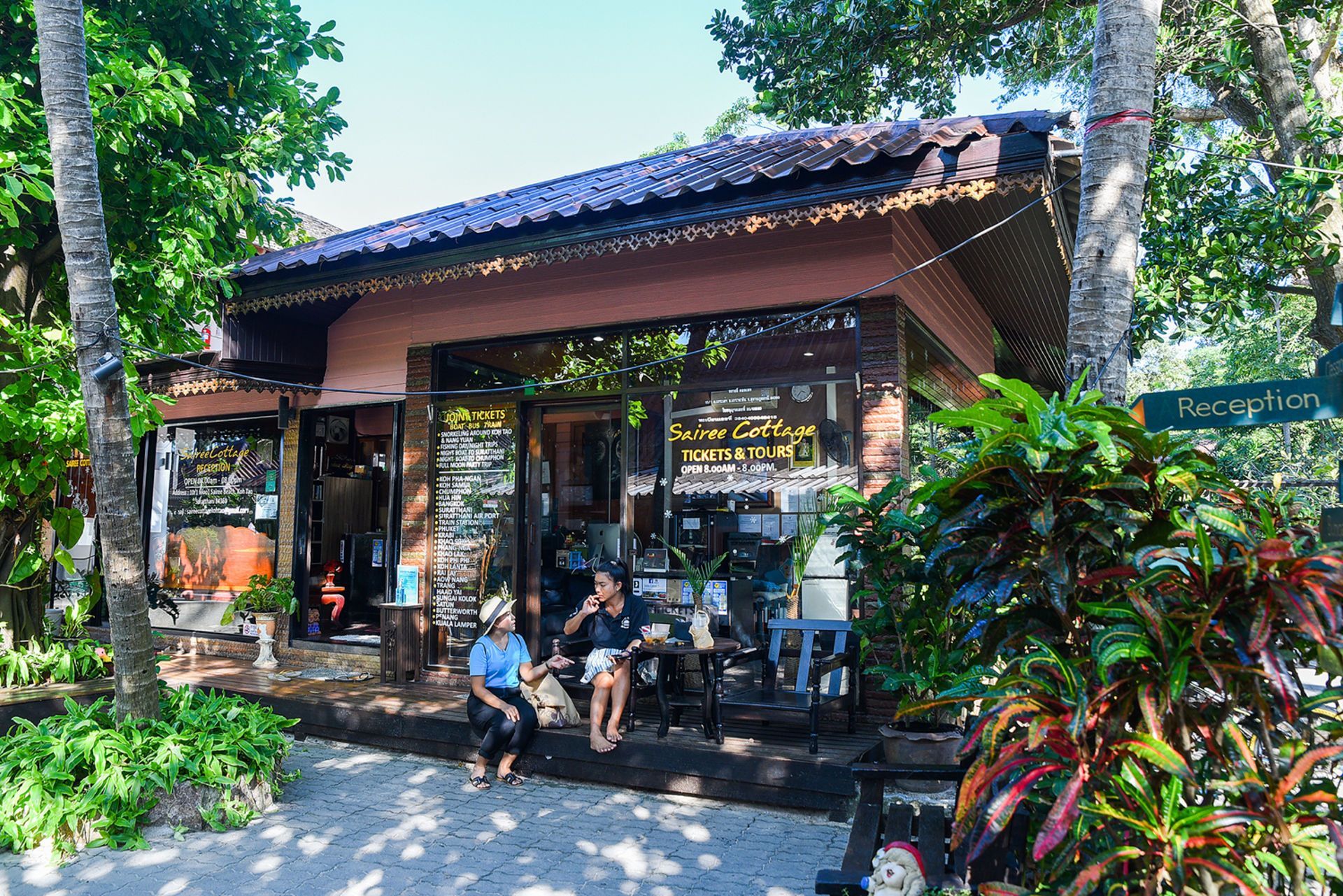 Shop with glass windows, two people sitting outside, lush greenery, and a tiled roof.