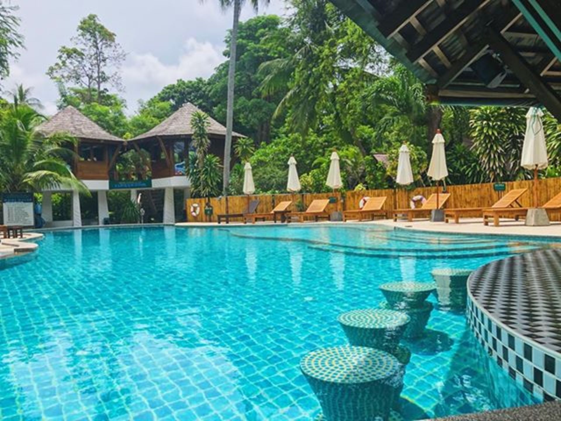 Swimming pool at a resort with pool-side bar, lounge chairs, and two elevated huts surrounded by lush greenery.