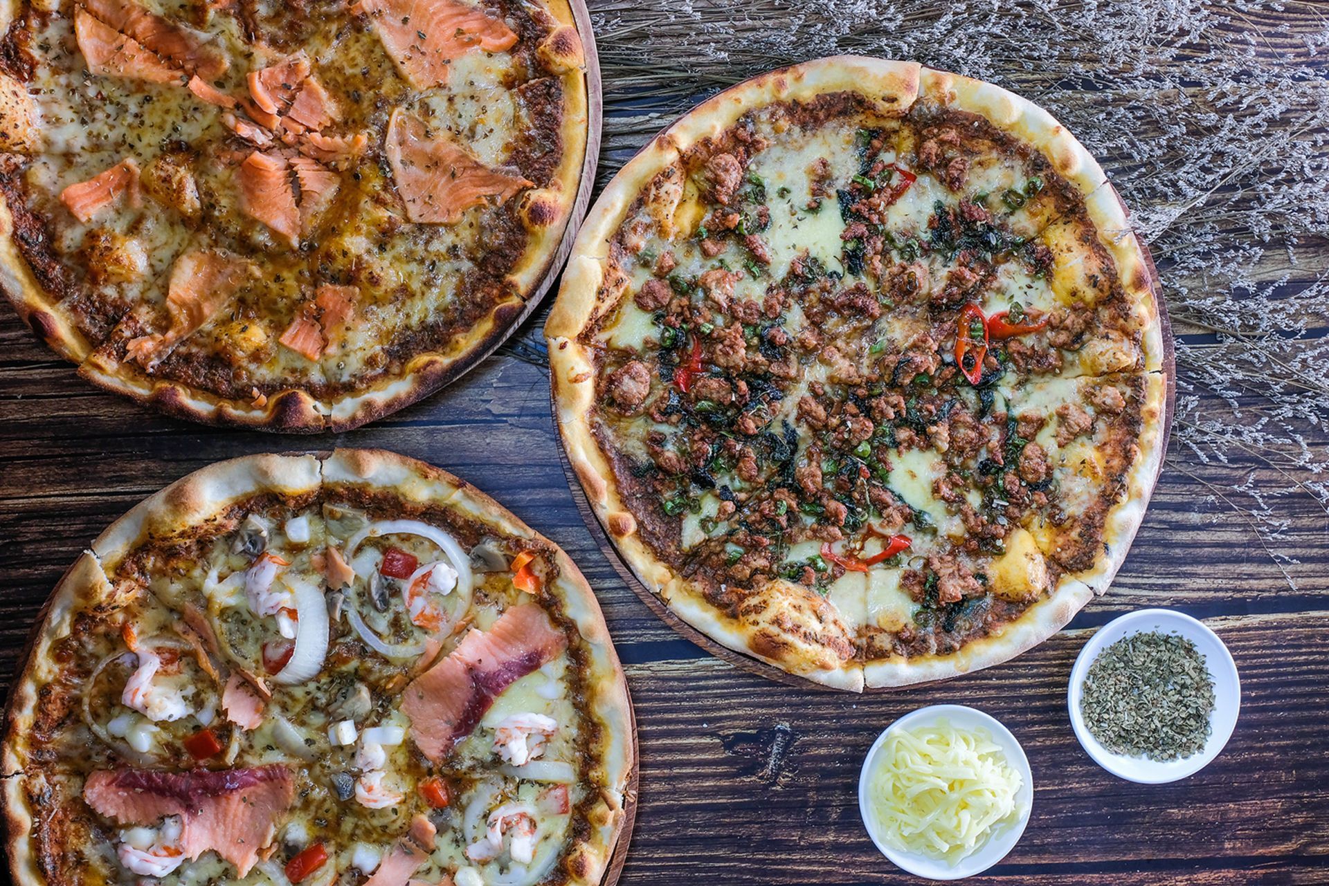 Three pizzas on a wooden table, with cheese and herbs in small white bowls.