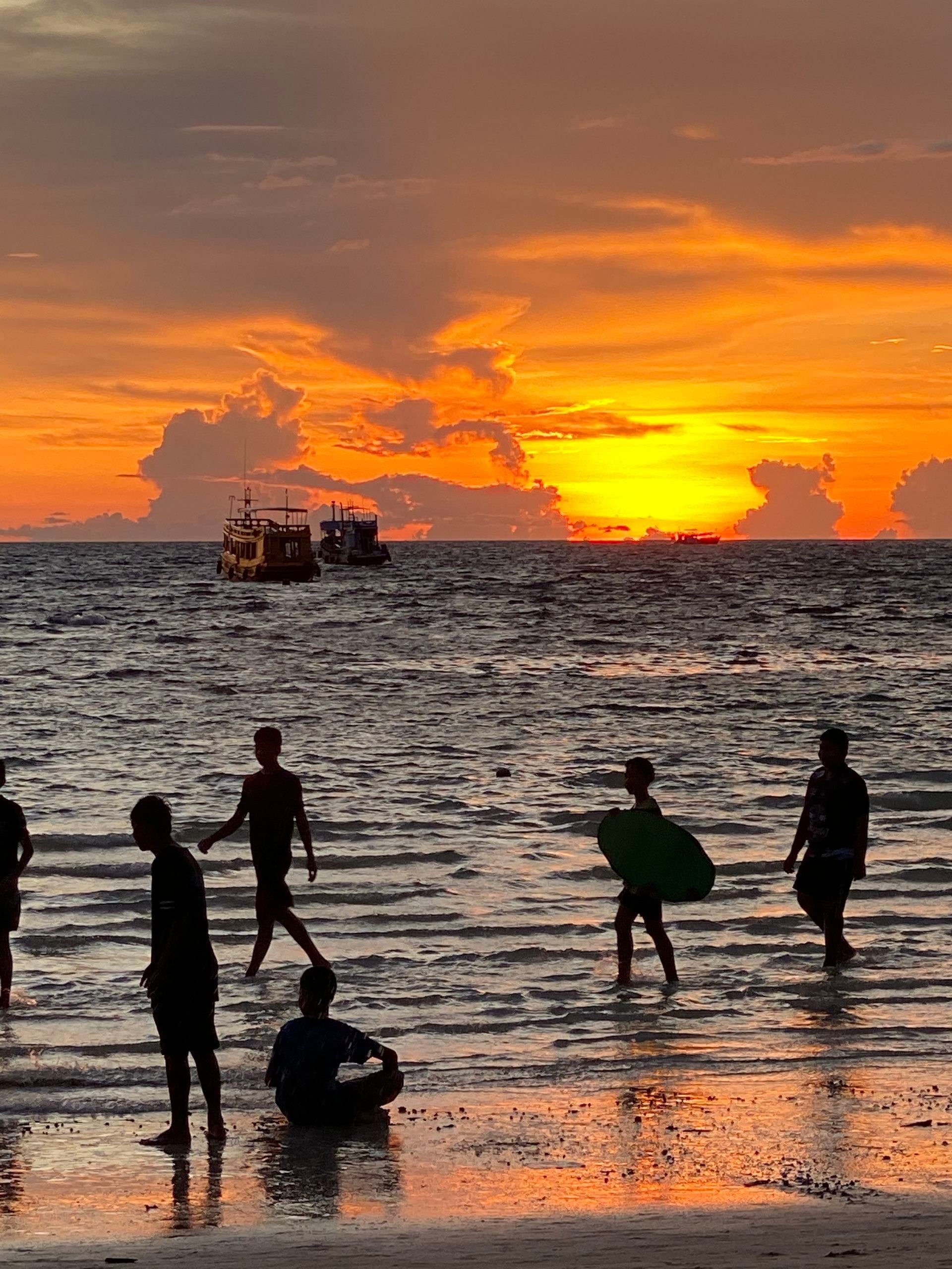 Silhouetted people wading in the ocean at sunset with orange and yellow sky, two boats in the distance.