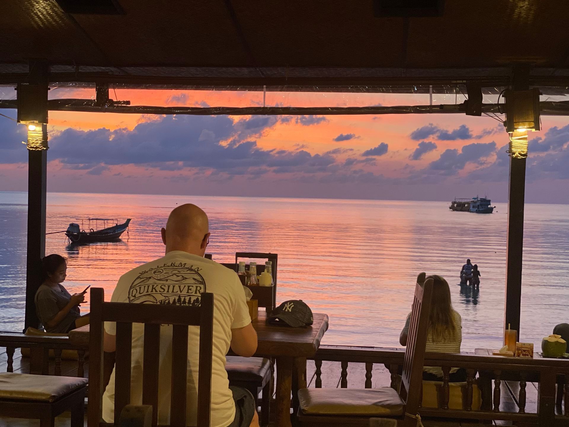 People dine at a restaurant overlooking the ocean at sunset; boats on the water, orange sky.