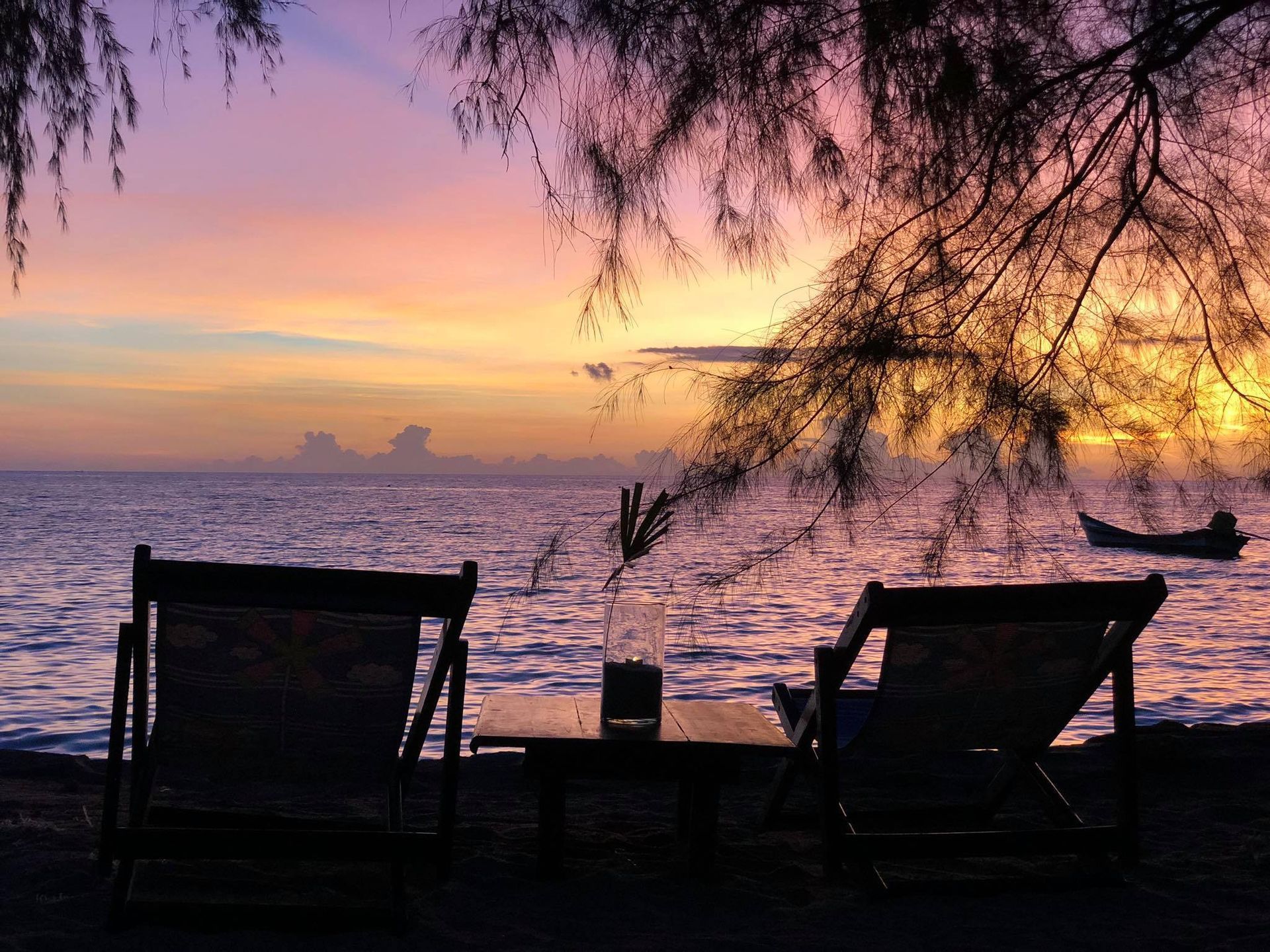 Two beach chairs face a sunset over the ocean. A small boat is visible.