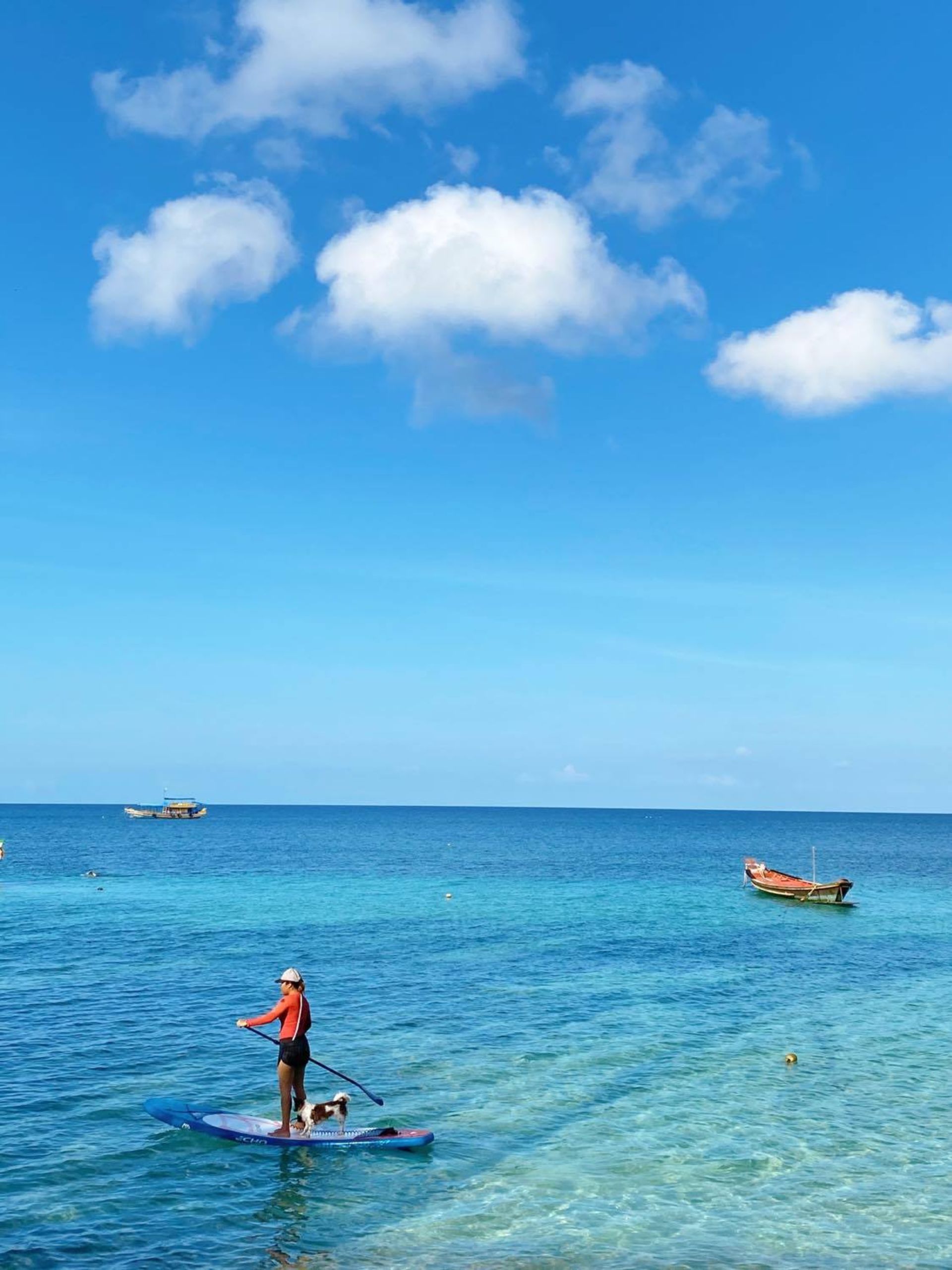 Person paddleboarding on a blue sea under a bright blue sky with fluffy white clouds, boats in distance.