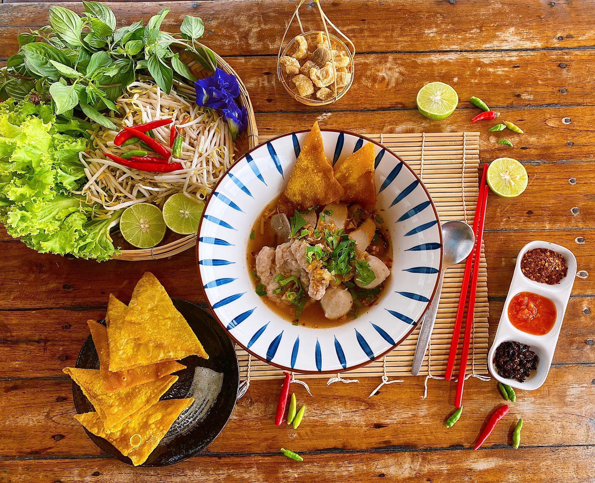 Bowl of noodles with garnishes on wooden table, includes vegetables, fried wontons, and chili.