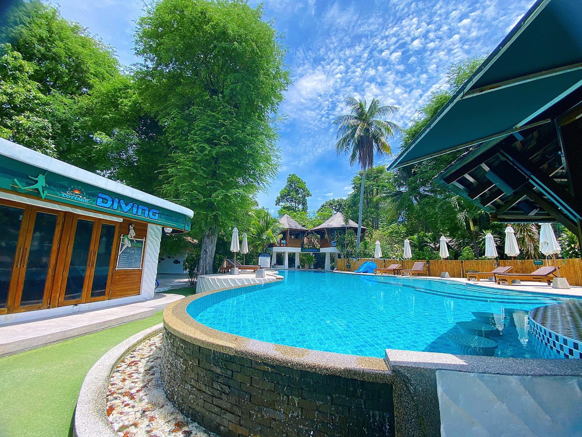 Swimming pool at a resort, turquoise water, wooden buildings, palm trees, sunny day.
