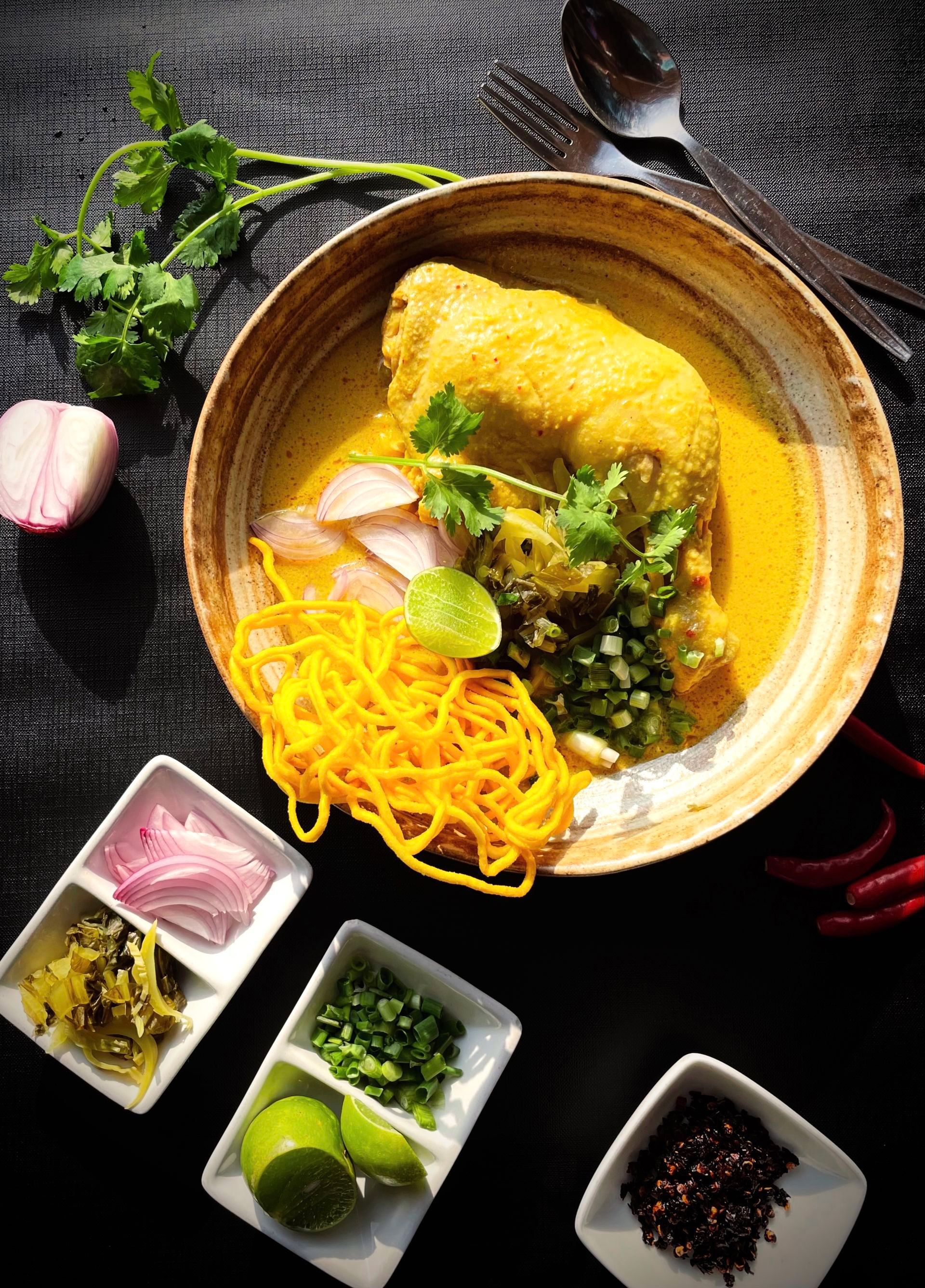 Thai khao soi dish with chicken, noodles, and condiments in a bowl on a dark surface.