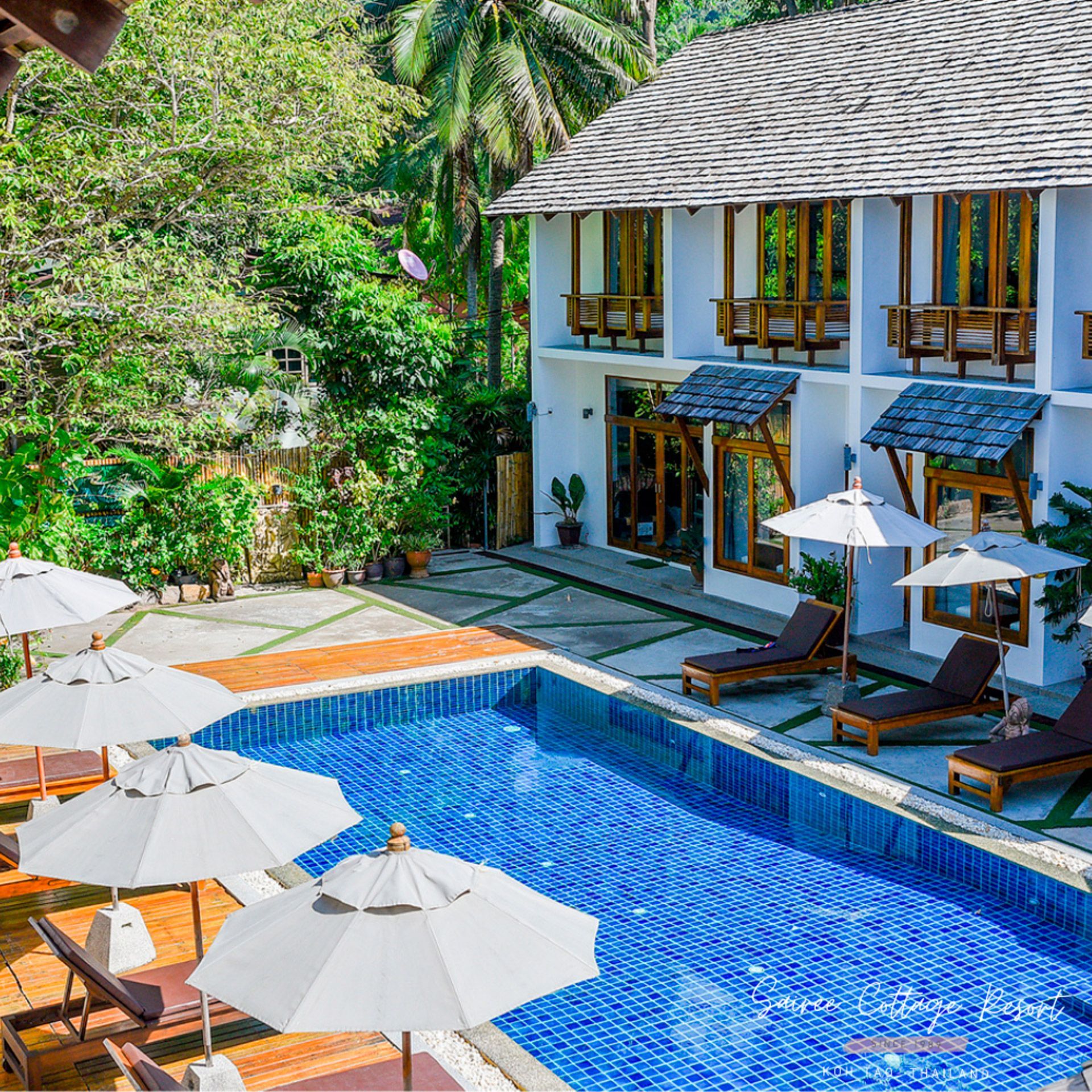 Swimming pool with sunbeds, white umbrellas, and a two-story white building in a tropical setting.
