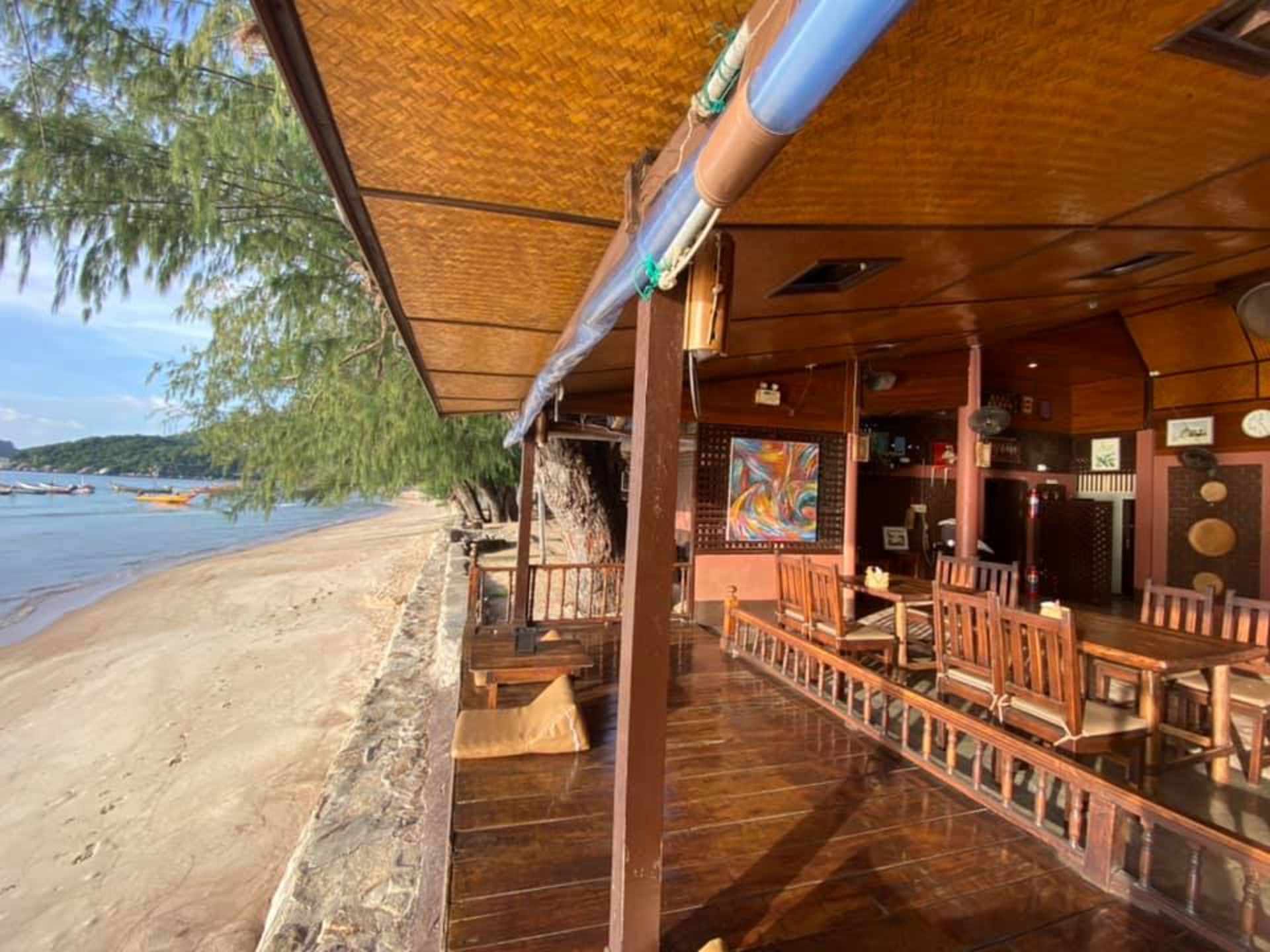 Beachside restaurant: wooden tables, thatched roof, sandy beach, boats in the distance.