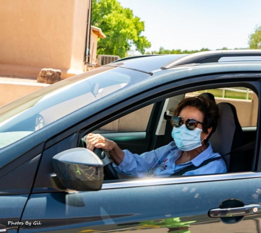 A woman wearing a mask and sunglasses drives a blue car on a sunny day. 