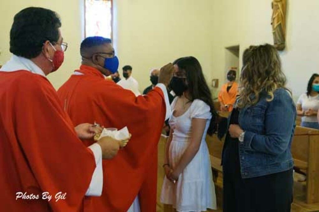 A priest in red robes blesses a young girl in a white dress during a church ceremony.