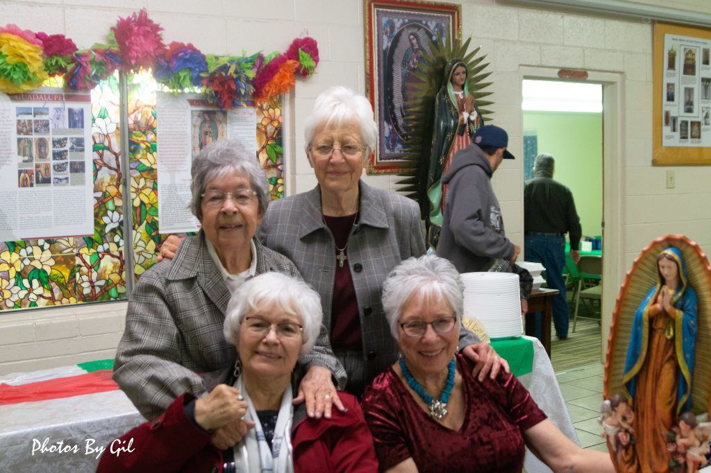 Four elderly women smile warmly in front of a colorful, festive backdrop with religious imagery.