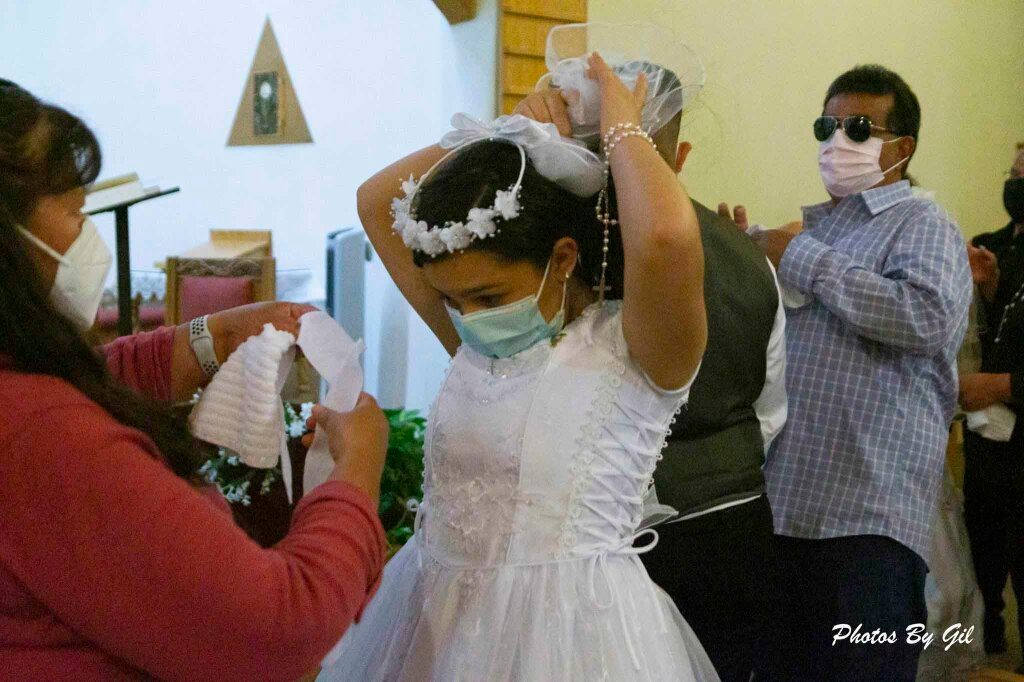 A young girl in a white dress, wearing a face mask, adjusts her veil with the help of a woman. 