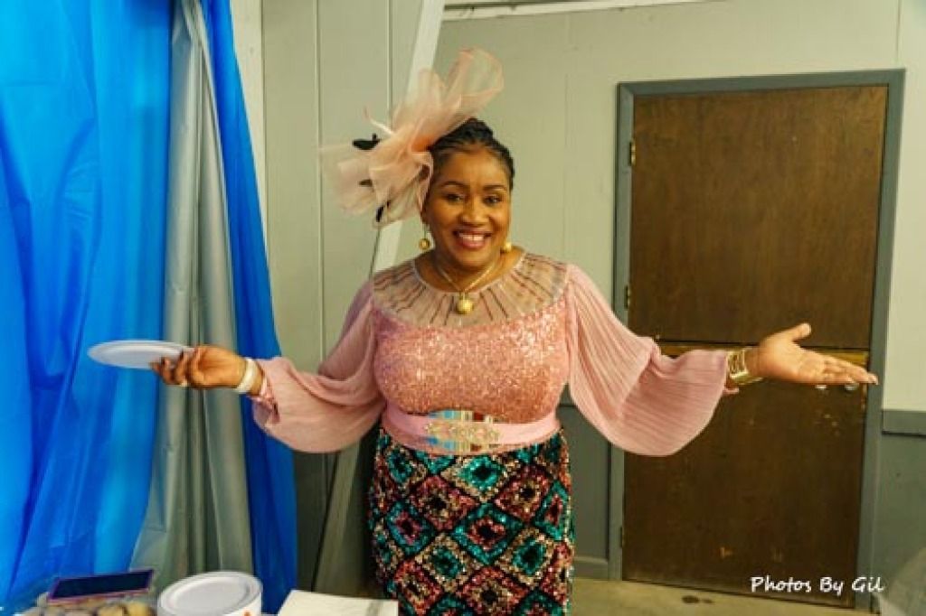A woman in a colorful, elegant outfit stands smiling with open arms. 