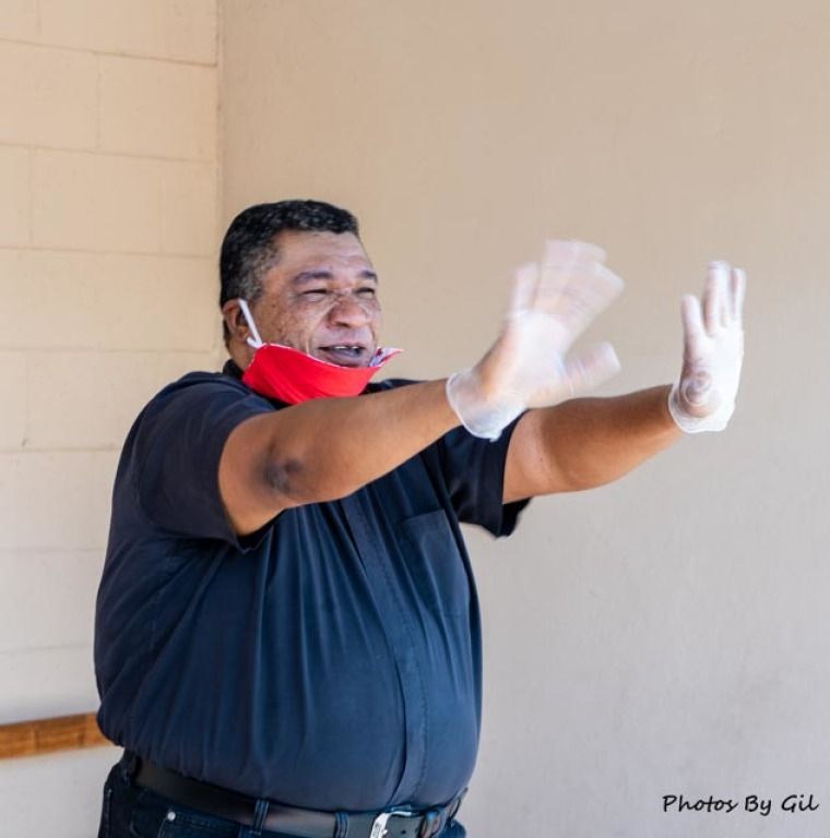 A smiling man wearing gloves and a partially lowered red mask gestures enthusiastically with open hands.