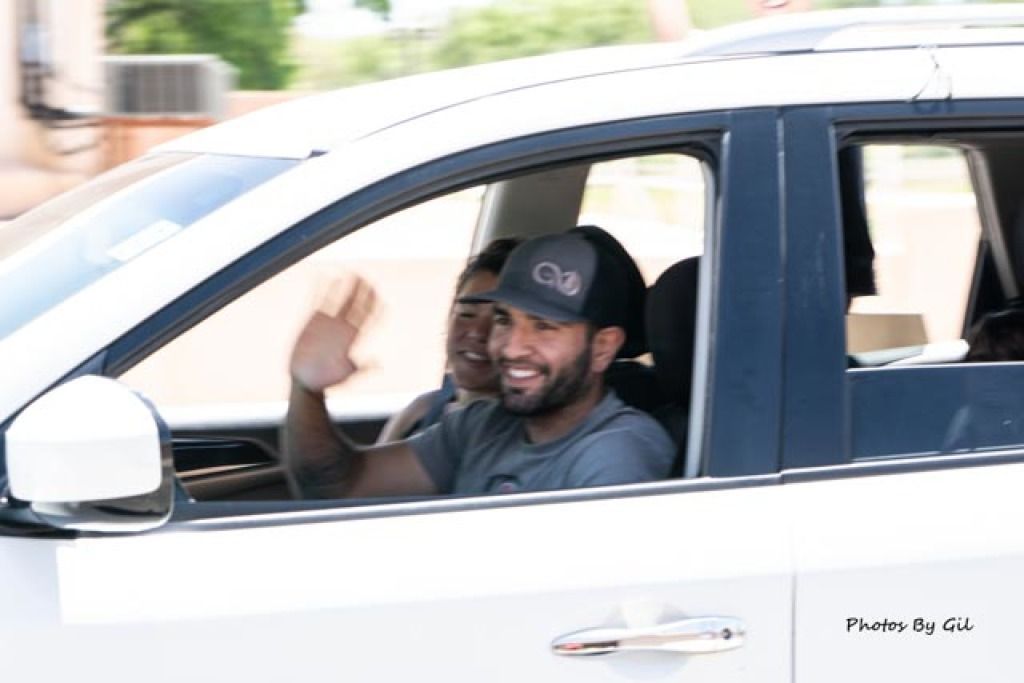 A smiling man in a gray cap waves from the driver's seat of a white car. 