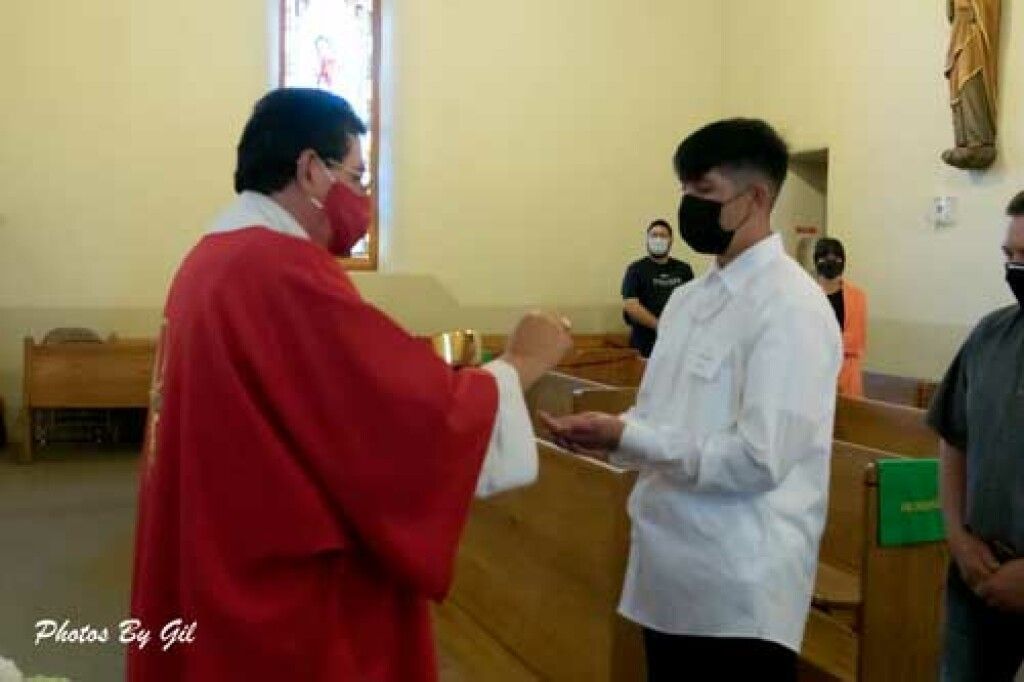 Priest in red robe giving communion to a masked young man in white, inside a church.