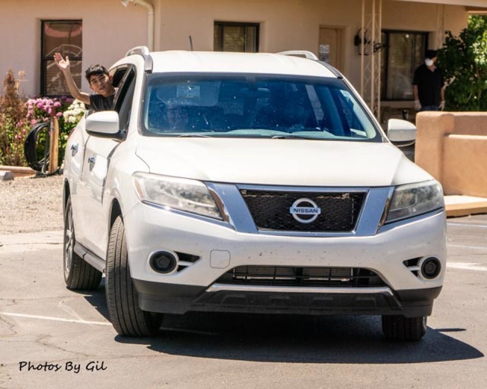 A person waves from the driver's window of a white Nissan SUV parked outside a building.
