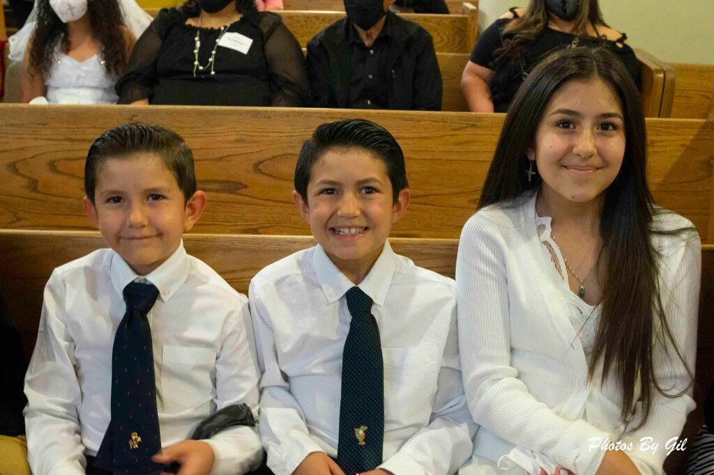 Three children dressed in white shirts sit smiling in a church pew. 