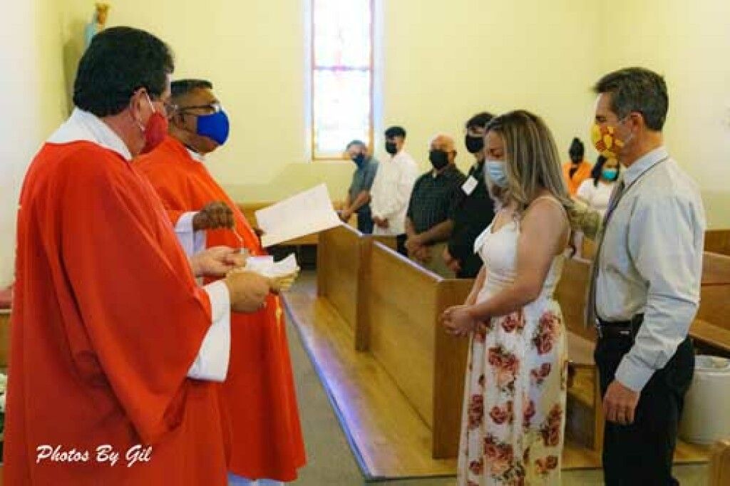 Two clergy in red vestments, wearing masks, face a couple dressed formally.