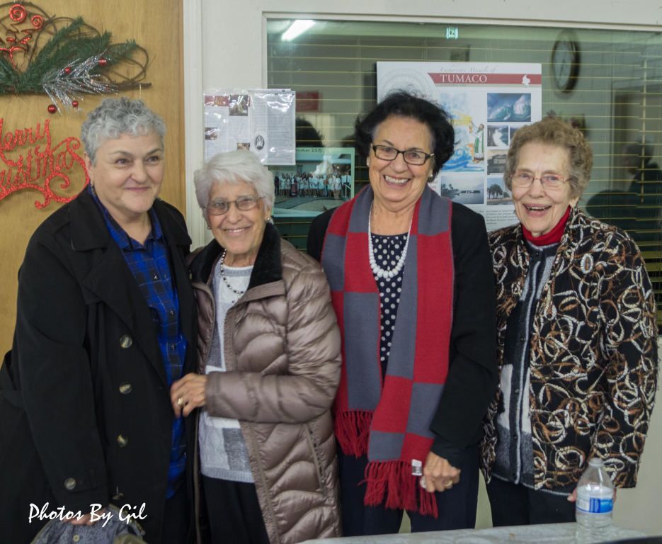Four smiling elderly women stand together indoors, dressed warmly.
