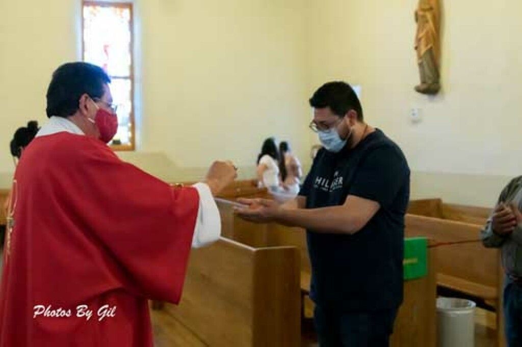 A man wearing a red vestment and mask offers communion to a masked churchgoer in a pew. 