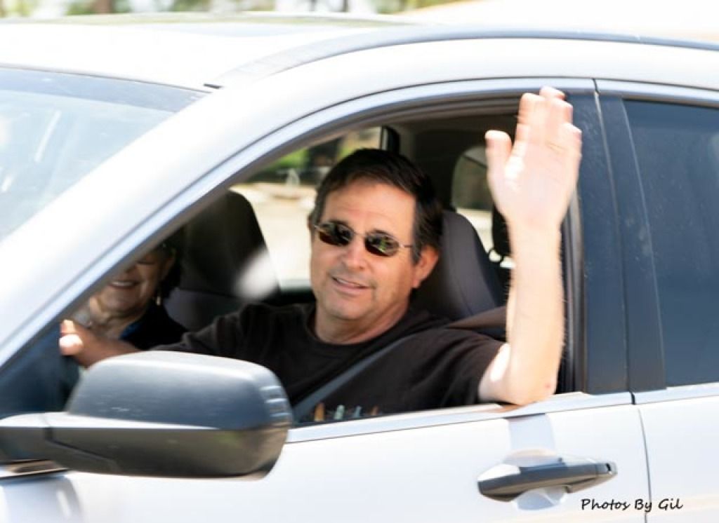 A man in sunglasses driving a car waves cheerfully out the window.