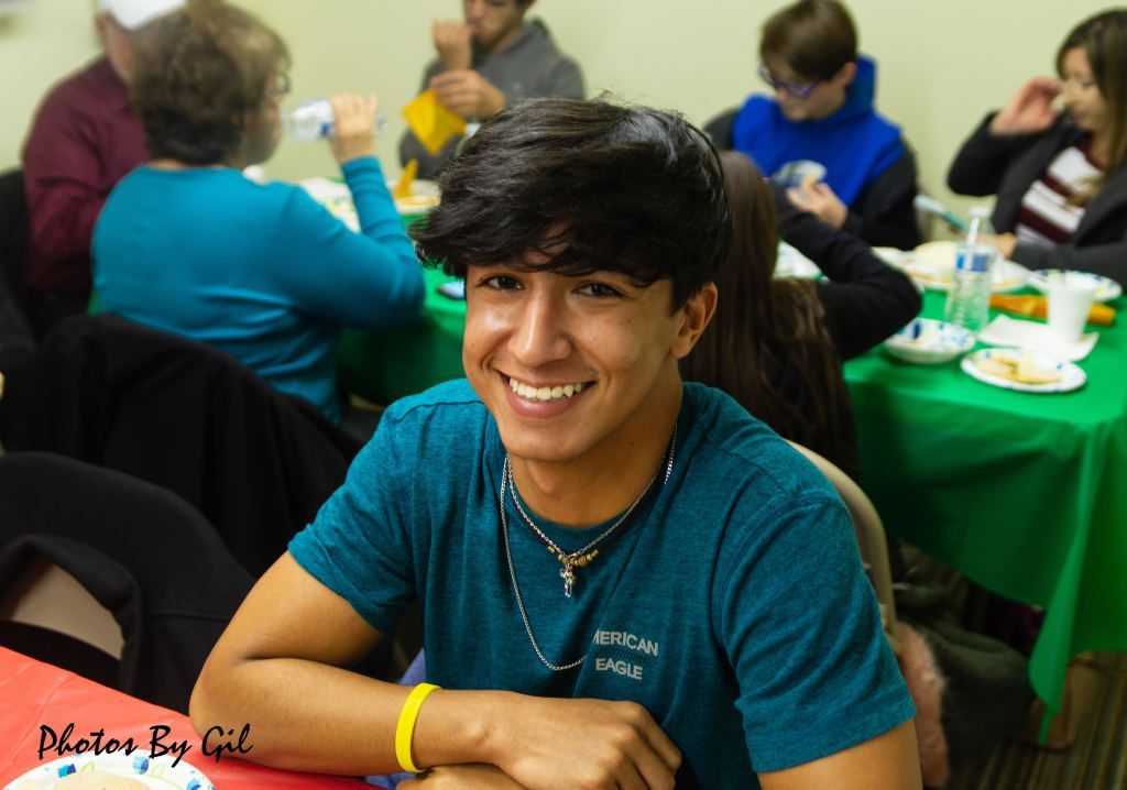 Smiling young man in a teal shirt sits at a table during a casual gathering.