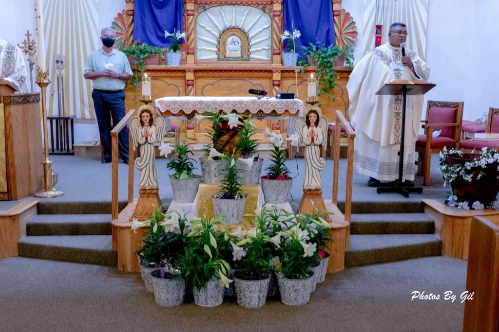 A priest in white robes speaks at a podium beside an altar adorned with angel statues and potted plants.