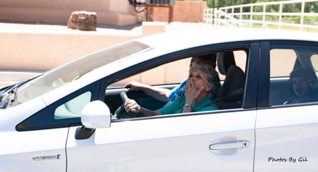 A white car with an older woman in the front seat, blowing a kiss with her hand. 