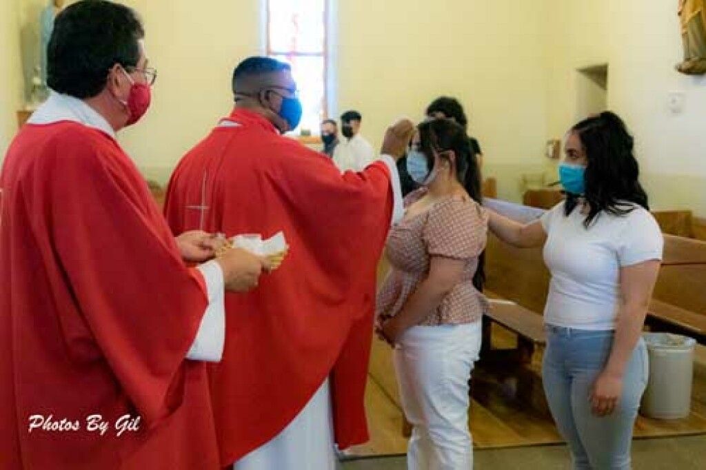 A priest in red vestments blesses a woman wearing a mask in a church. 
