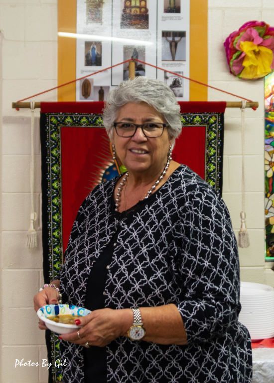 Elderly woman with short gray hair, wearing glasses and a patterned shirt, smiles while holding a bowl. 