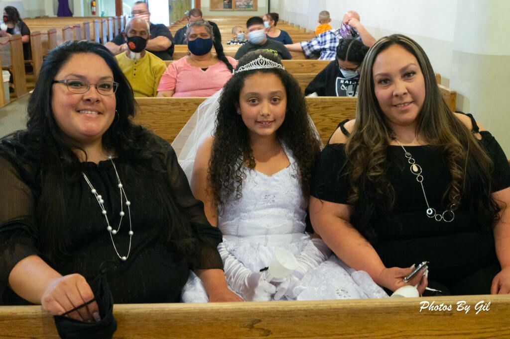 A young girl in a white dress and tiara is between two women in black. 