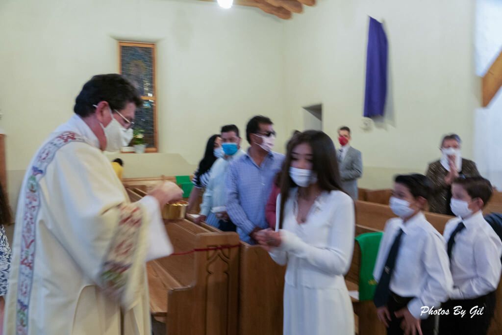 A priest in a cream-colored robe distributes communion to a young woman in white. 