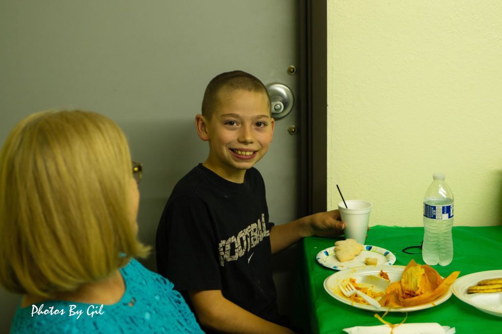A smiling boy in a black shirt sits at a table with plates of food and a water bottle. 