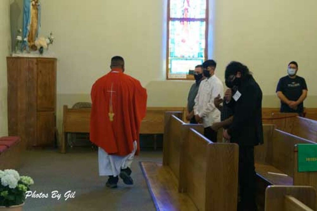 A priest in red vestments blesses four masked men inside a church with wooden pews.