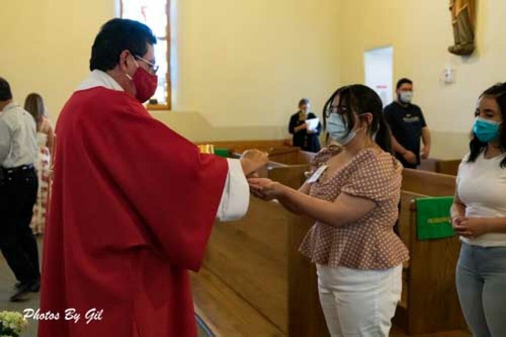 A priest in a red robe, wearing a mask, distributes communion to a masked woman in a church.
