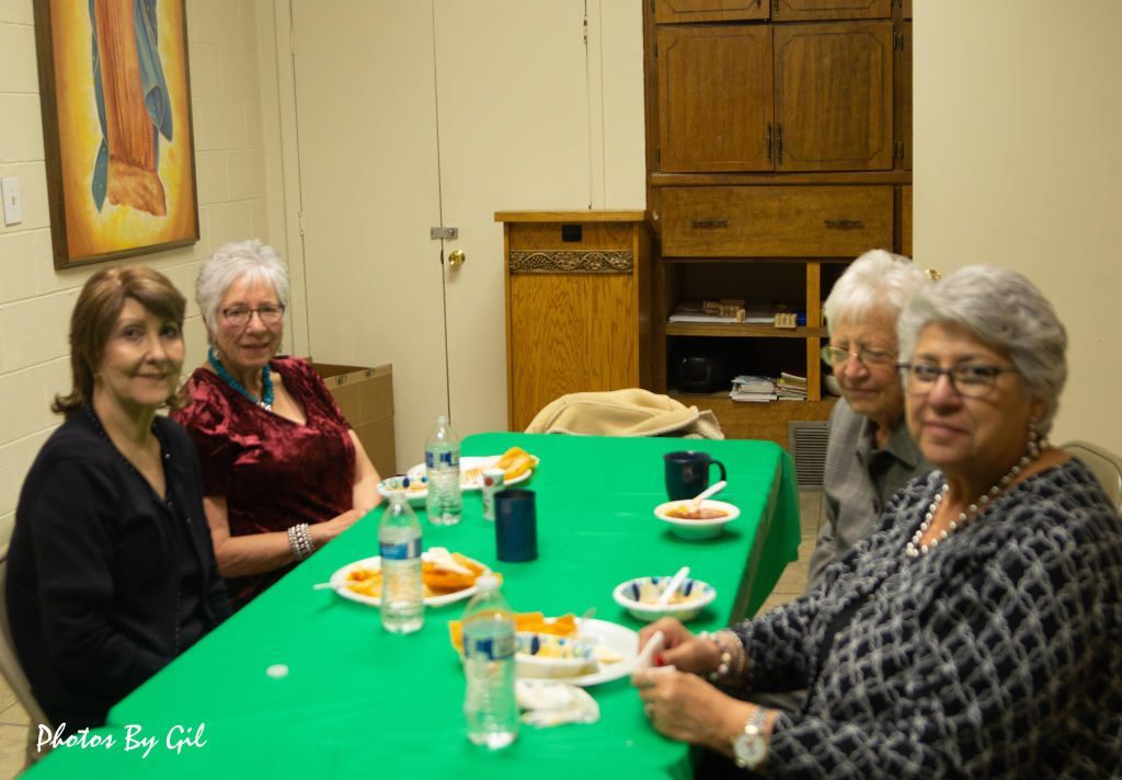 Four women sit at a green tablecloth, enjoying snacks and drinks, in a cozy room.